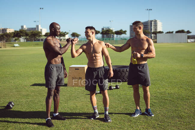 Diverse group of happy shirtless men exercising outdoors, taking a break talking and bumping fists. healthy active lifestyle, cross training for fitness. — Stock Photo