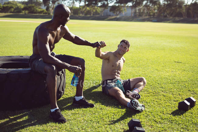 Two happy shirtless men exercising outdoors, taking a break talking and bumping fists. healthy active lifestyle, cross training for fitness. — Stock Photo