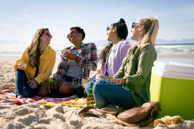 Счастливая группа разнообразных подруг, веселящихся, сидящих на пляже и смеющихся. отдых, свобода и досуг на открытом воздухе. — стоковое фото