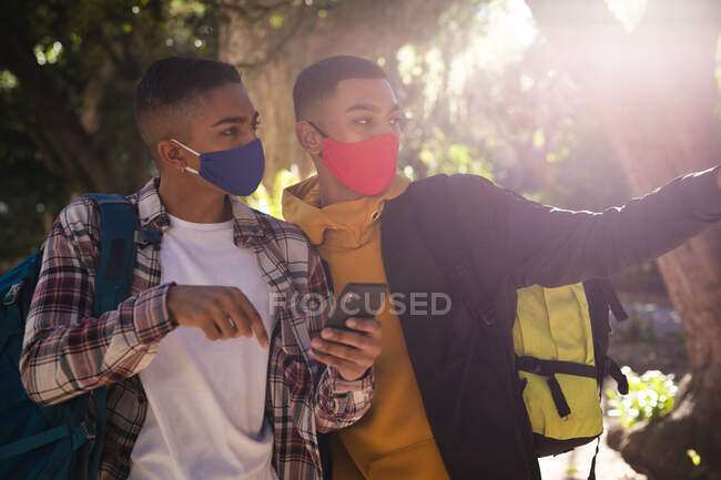 Zwei gemischte Rassenfreunde mit Gesichtsmasken und Rucksäcken im Stadtpark per Smartphone. Rucksackurlaub, Städtereise während der Coronavirus-Pandemie. — Stockfoto