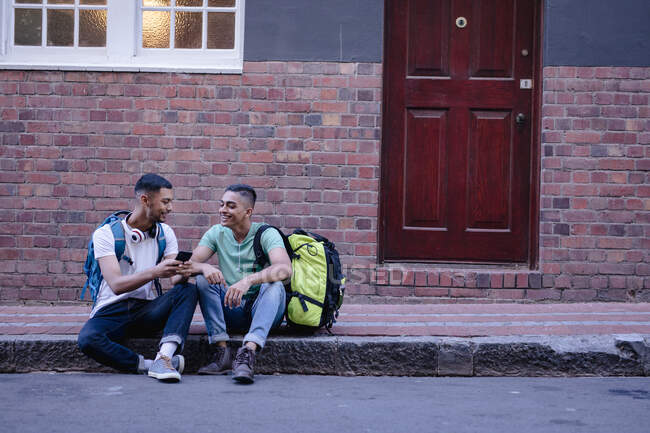 Two happy mixed race male friends with backpacks sitting in city street talking, using smartphone. backpacking holiday, city travel break. — Stock Photo