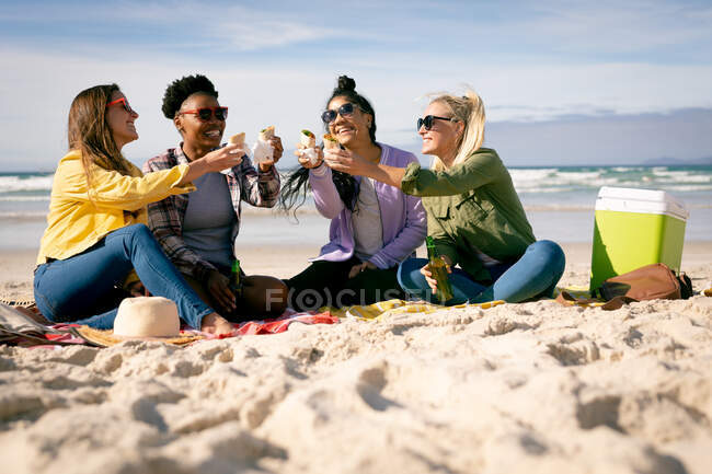 Happy group of diverse female friends having fun, siting on beach holding food laughing. holiday, freedom and leisure time outdoors. — Stock Photo