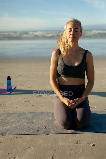 Mujer caucásica practicando yoga, de rodillas meditando en la playa. estilo de vida activo saludable, fitness al aire libre y bienestar. - foto de stock