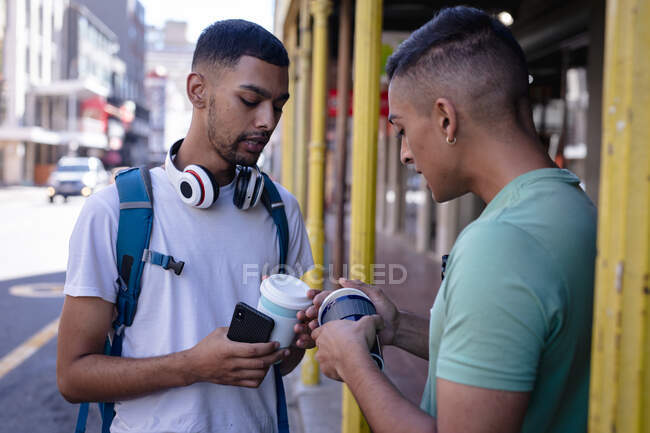Dos amigos varones de raza mixta con mochilas de pie en la calle de la ciudad con cafés para llevar, hablando. vacaciones de mochilero, escapada a la ciudad. - foto de stock