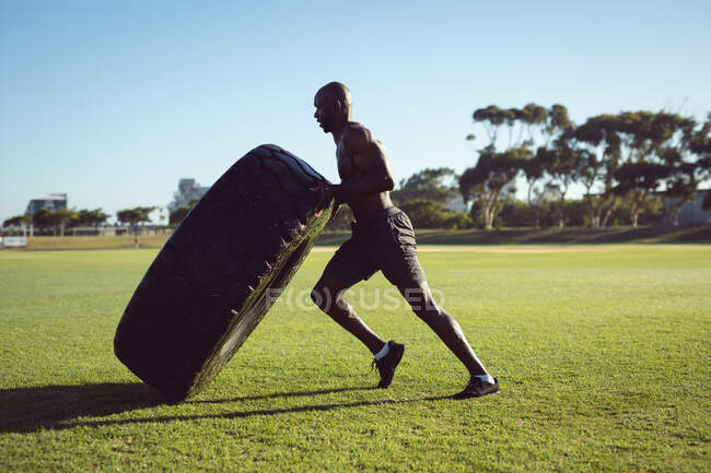 Un hombre afroamericano en forma de camisa haciendo ejercicio al aire libre, levantando neumáticos pesados. estilo de vida activo saludable, entrenamiento cruzado para fitness. - foto de stock