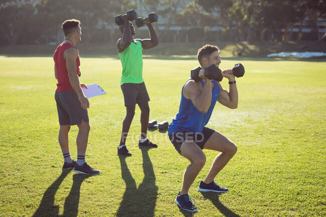 Zwei fitte Männer und Trainer trainieren im Freien und heben Hanteln. gesunder aktiver Lebensstil, Crosstraining für Fitness. — Stockfoto