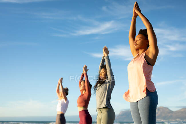 Gruppo di diverse amiche che praticano yoga, in piedi e alzando le mani sulla spiaggia. sano stile di vita attivo, fitness e benessere all'aperto. — Foto stock