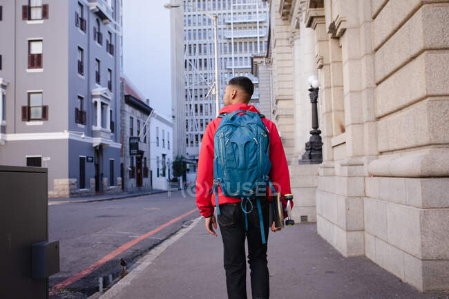 Vista trasera del hombre de raza mixta que lleva mochila y monopatín caminando en la calle de la ciudad. vacaciones de mochilero, escapada a la ciudad. - foto de stock