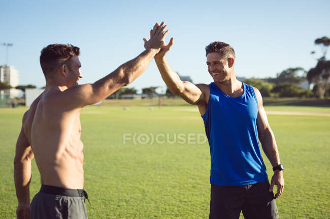 Два счастливых кавказских мускулистых человека, тренирующихся на улице, улыбающихся и дающих пять. здоровый активный образ жизни, кросс тренировки для фитнеса. — стоковое фото