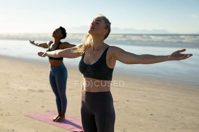 Две разноплановые женщины практикуют йогу, стоя с протянутыми руками, медитируя на пляже. здоровый активный образ жизни, фитнес на открытом воздухе и благополучие. — стоковое фото