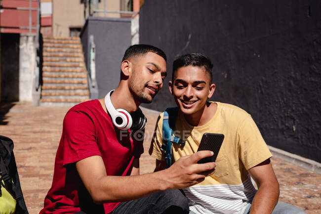 Dos sonrientes amigos varones de raza mixta sentados en la soleada calle de la ciudad tomando selfie con smartphone. vacaciones de mochilero, escapada a la ciudad. - foto de stock
