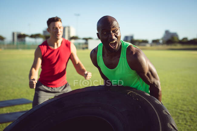 Hombre y entrenador en forma diversa haciendo ejercicio al aire libre, alentando y levantando neumáticos pesados. estilo de vida activo saludable, entrenamiento cruzado para fitness. - foto de stock