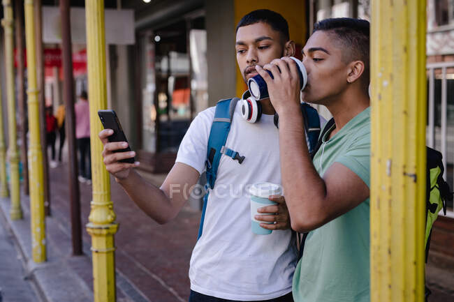 Due amici maschi di razza mista con zaini in piedi in città a bere caffè, utilizzando lo smartphone. vacanza zaino in spalla, city travel break. — Foto stock