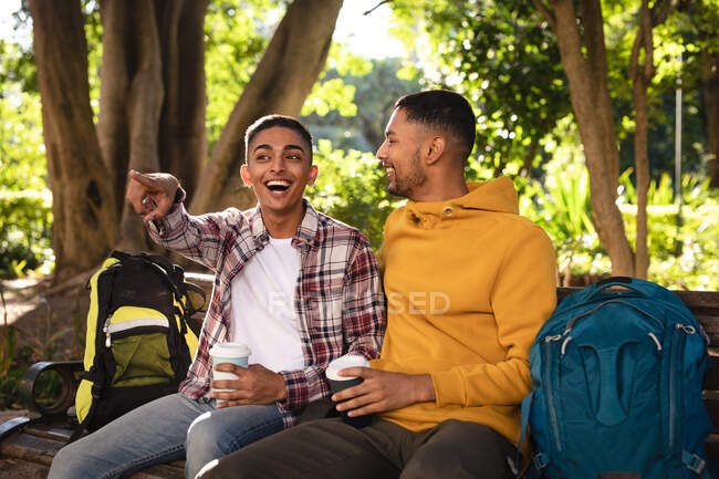 Dos felices amigos varones de raza mixta sentados en el banco del parque con mochilas, hablando. vacaciones de mochilero, escapada a la ciudad. - foto de stock