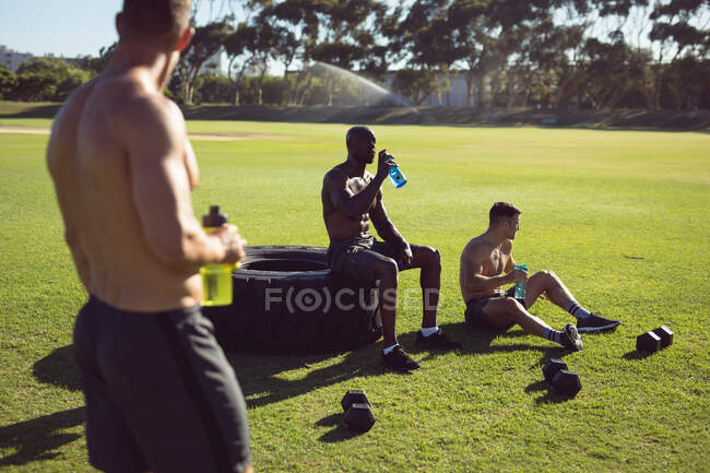 Diverso gruppo di uomini felici senza maglietta che si esercitano all'aperto, facendo una pausa parlando e bevendo acqua. sano stile di vita attivo, cross training per il fitness. — Foto stock