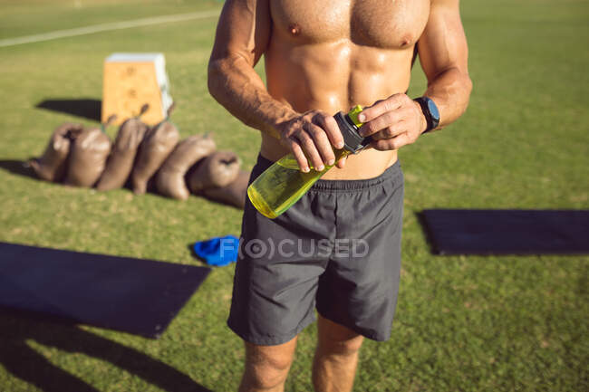 Mid section muscular shirtless man drinking water taking a break during exercise outdoors. healthy active lifestyle, cross training for fitness. — Stock Photo