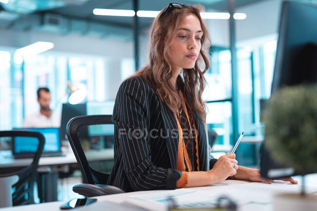 Mujer de negocios caucásica sentado en la mesa y el uso de la computadora con colegas en segundo plano. trabajar en una oficina moderna. - foto de stock