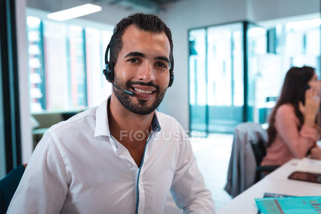 Портрет змішаної раси бізнесмен в гарнітурі і сидить за столом посміхаючись. робота в сучасному офісі . — стокове фото