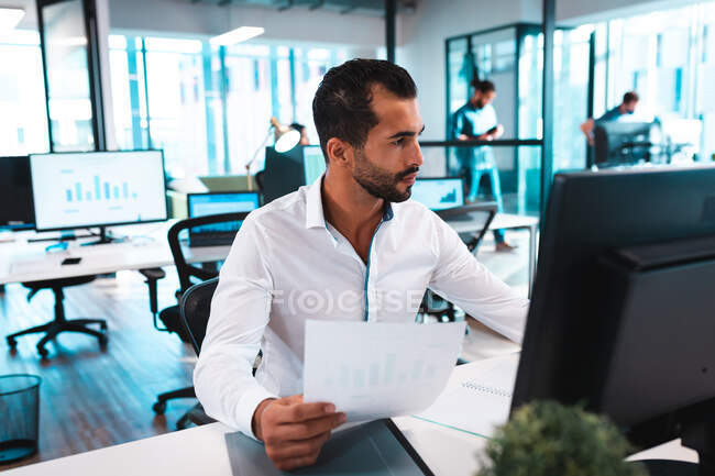 Empresário de raça mista à mesa segurando documentos e usando computador com colegas em segundo plano. trabalho em um escritório moderno. — Fotografia de Stock