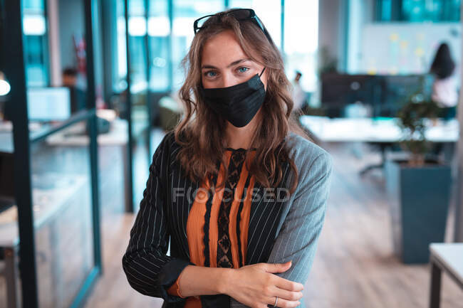 Retrato de mujer de negocios caucásica con máscara facial con colegas de fondo. trabajar en una oficina moderna durante la pandemia de coronavirus covid 19. - foto de stock