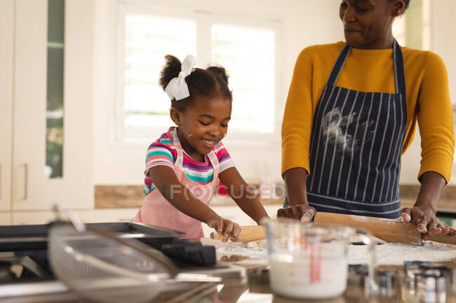 Улыбающиеся африканская американская мать и дочь пекут на кухне скручивая тесто вместе. Семья проводит время вместе дома. — стоковое фото