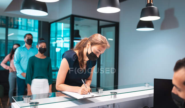 Grupo de empresarios diversos que llevan máscaras faciales y se inscriben en la recepción. trabajar en una oficina moderna durante la pandemia de coronavirus covid 19. - foto de stock