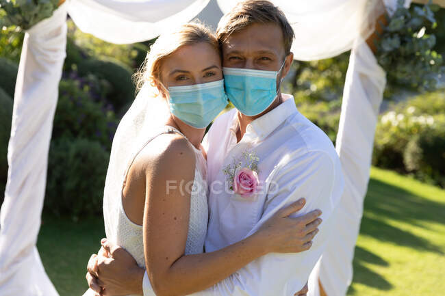Portrait of happy caucasian bride and groom getting married wearing face masks and embracing. summer wedding, marriage, love and celebration during covid 19 pandemic concept. — Stock Photo