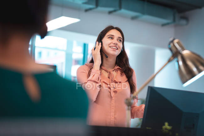 Caucasian businesswoman wearing headset and sitting at table talking, using computer. work at a modern office. — Stock Photo
