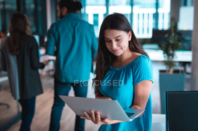 Mixed race businesswoman smiling and using laptop with colleagues in background. work at a modern office. — Stock Photo