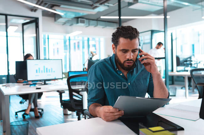 Mixed race businessman sitting at table using smartphone with colleagues in background. work at a modern office. — Stock Photo
