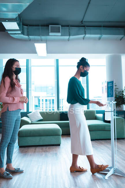 Two diverse businesswomen wearing face masks and disinfecting hands at work. work at a modern office during covid 19 coronavirus pandemic. — Stock Photo