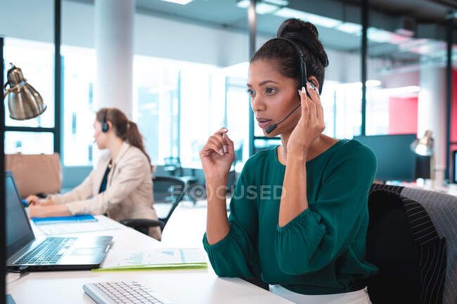 Donna d'affari mista che indossa auricolari e si siede a tavola a parlare, usando il computer. lavorare in un ufficio moderno. — Foto stock