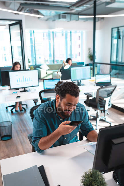 Empresario de carrera mixta sentado en la mesa usando smartphone con colegas en segundo plano. trabajar en una oficina moderna. - foto de stock