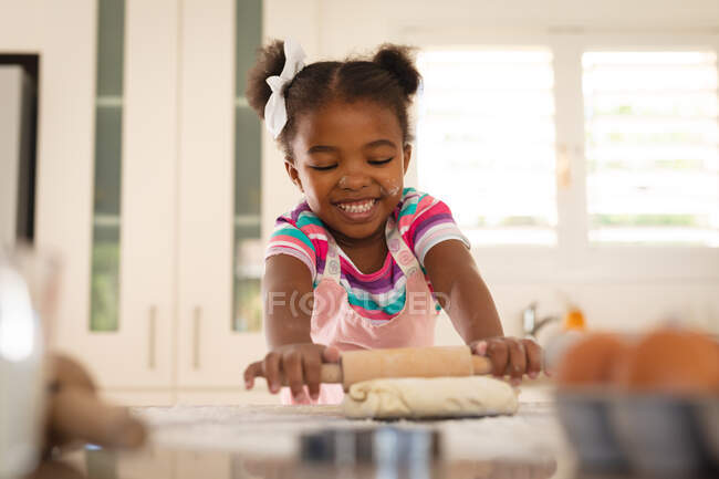 Счастливая африканская американская дочь, опирающаяся на кухонный стол, скручивая тесто скалкой. проводить свободное время дома. — стоковое фото