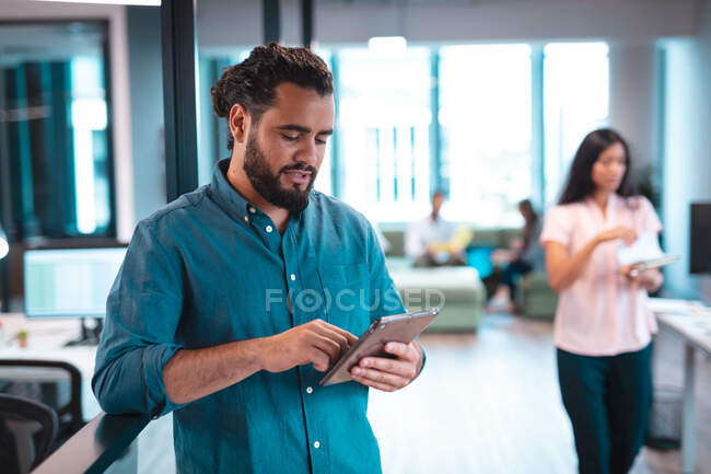 Homme d'affaires mixte utilisant une tablette avec des collègues travaillant en arrière-plan. travailler dans un bureau moderne. — Photo de stock