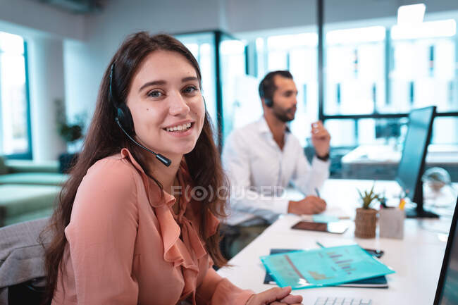 Портрет кавказской предпринимательницы в наушниках и сидящей за столом. работа в современном офисе. — стоковое фото