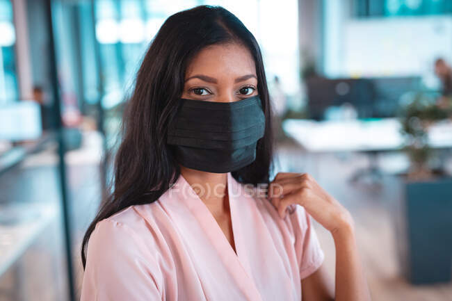 Retrato de una mujer de negocios de raza mixta que usa mascarilla facial con colegas de fondo. trabajar en una oficina moderna durante la pandemia de coronavirus covid 19. - foto de stock