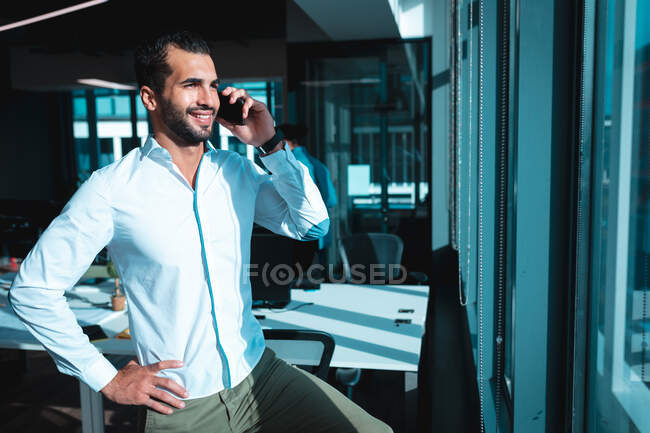 Homme d'affaires mixte à fenêtre parlant sur smartphone avec des collègues en arrière-plan. travailler dans un bureau moderne. — Photo de stock