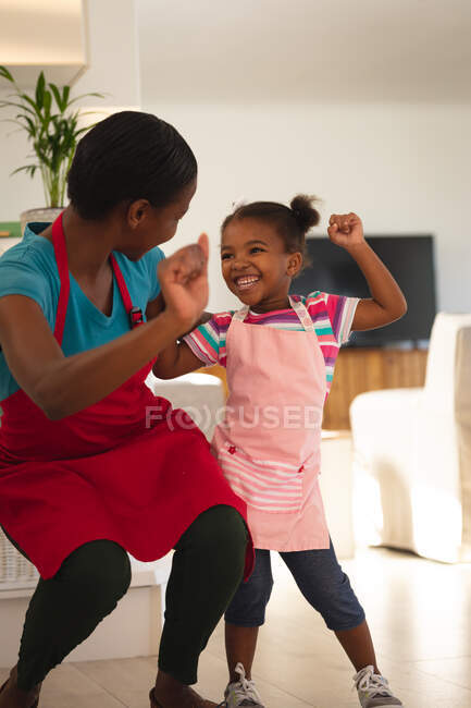 Happy african american mother and daughter baking in kitchen doing celebration dance wearing aprons. family spending time together at home. — Stock Photo