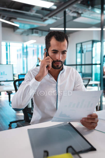 Empresário de raça mista à mesa segurando documentos e conversando no smartphone. trabalho em um escritório moderno. — Fotografia de Stock