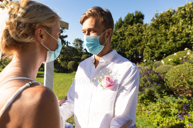 Heureux mariés caucasiens se marier portant des masques et se tenant la main. mariage d'été, mariage, amour et célébration pendant covid 19 concept de pandémie. — Photo de stock