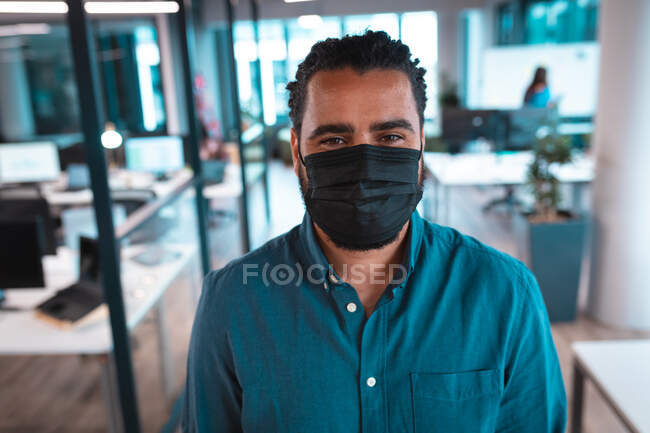 Portrait of mixed race businessman wearing face mask with colleagues in background. work at a modern office during covid 19 coronavirus pandemic. — Stock Photo