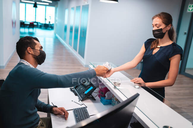 Dos empresarios diversos que usan máscaras faciales y toman temperatura en la recepción. trabajar en una oficina moderna durante la pandemia de coronavirus covid 19. - foto de stock