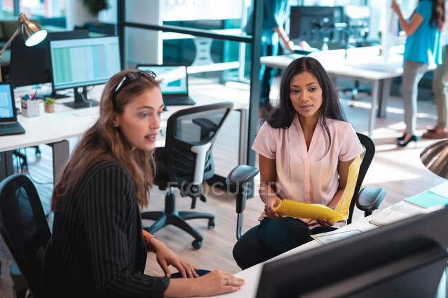 Duas mulheres de negócios diversas sentadas à mesa e usando computador com colegas em segundo plano. trabalho em um escritório moderno. — Fotografia de Stock