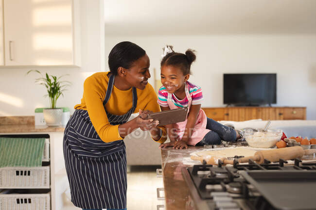 Sorrindo afro-americana mãe e filha assar na cozinha olhando receita em comprimido. família passar tempo juntos em casa. — Fotografia de Stock