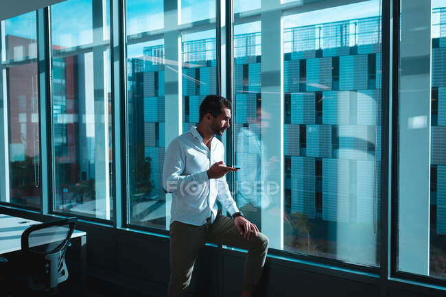 Empresário de corrida mista na janela usando smartphone e olhando para longe. trabalho em um escritório moderno. — Fotografia de Stock
