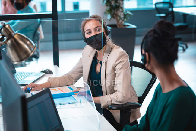 Dos empresarias diversas que usan mascarilla facial, hablando y usando computadora. trabajar en una oficina moderna durante la pandemia de coronavirus covid 19. - foto de stock
