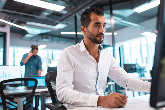 Geschäftsmann mit gemischter Rasse sitzt am Tisch und bedient den Computer mit Kollegen im Hintergrund. Arbeit in einem modernen Büro. — Stockfoto