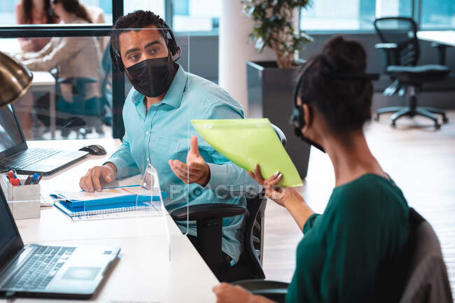Dois empresários diversos usando máscara facial, segurando documentos e usando o computador. trabalho em um escritório moderno durante covid 19 coronavirus pandemia. — Fotografia de Stock