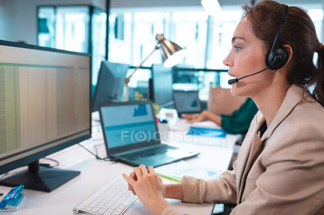 Mulher de negócios caucasiana usando fone de ouvido conversando e usando computador com colegas de fundo. trabalho em um escritório moderno. — Fotografia de Stock